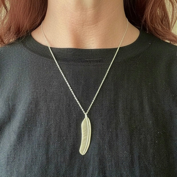 Person wearing a necklace with a feather shaped pendant on a plain background