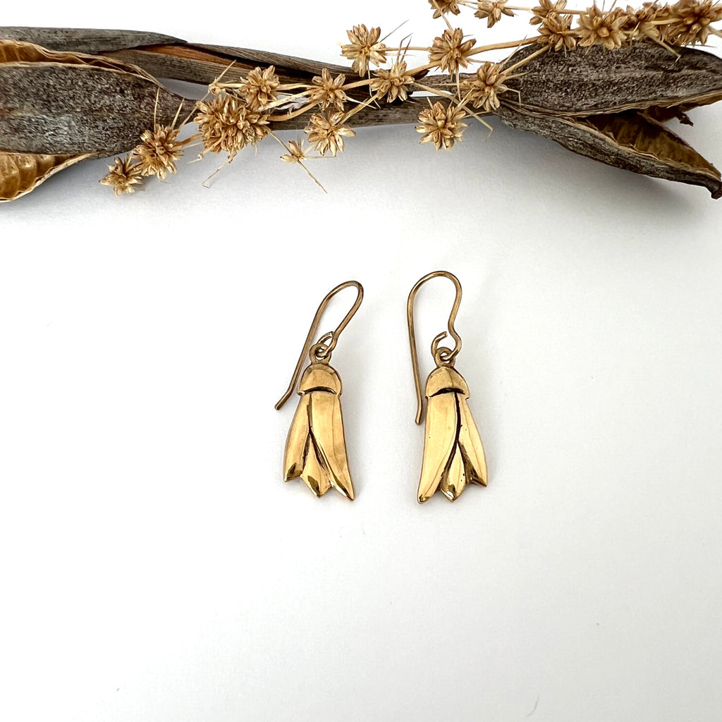 A pair of gold plated K\u010whai flower earrings on a white background, with the design inspired by the New Zealand native flower, placed next to some dried plant material.