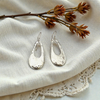 Silver earrings on a textured fabric surface with a branch and flowers.