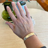 Hand wearing a gold bracelet on a wooden surface with a plant in the background