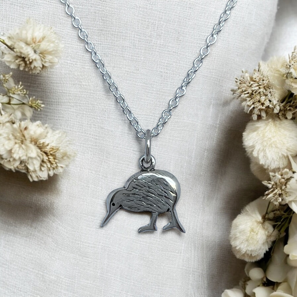 A sterling silver necklace featuring a petite kiwi bird pendant, displayed with a silver chain and clasp, against a background with dried flowers.