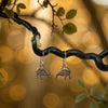 Pair of silver kiwi bird earrings hanging from a branch with a blurred bokeh background.