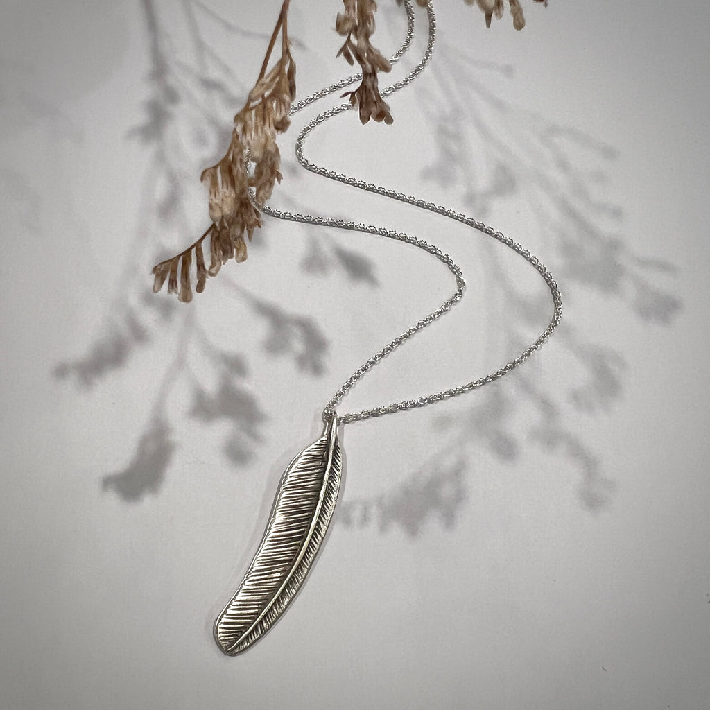 Silver feather necklace on a light gray background with dried leaves.