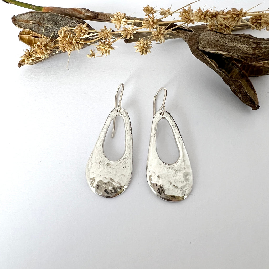 A pair of sterling silver freeform droplet earrings with a hammered texture, displayed against a white background with some dried plants to the side.