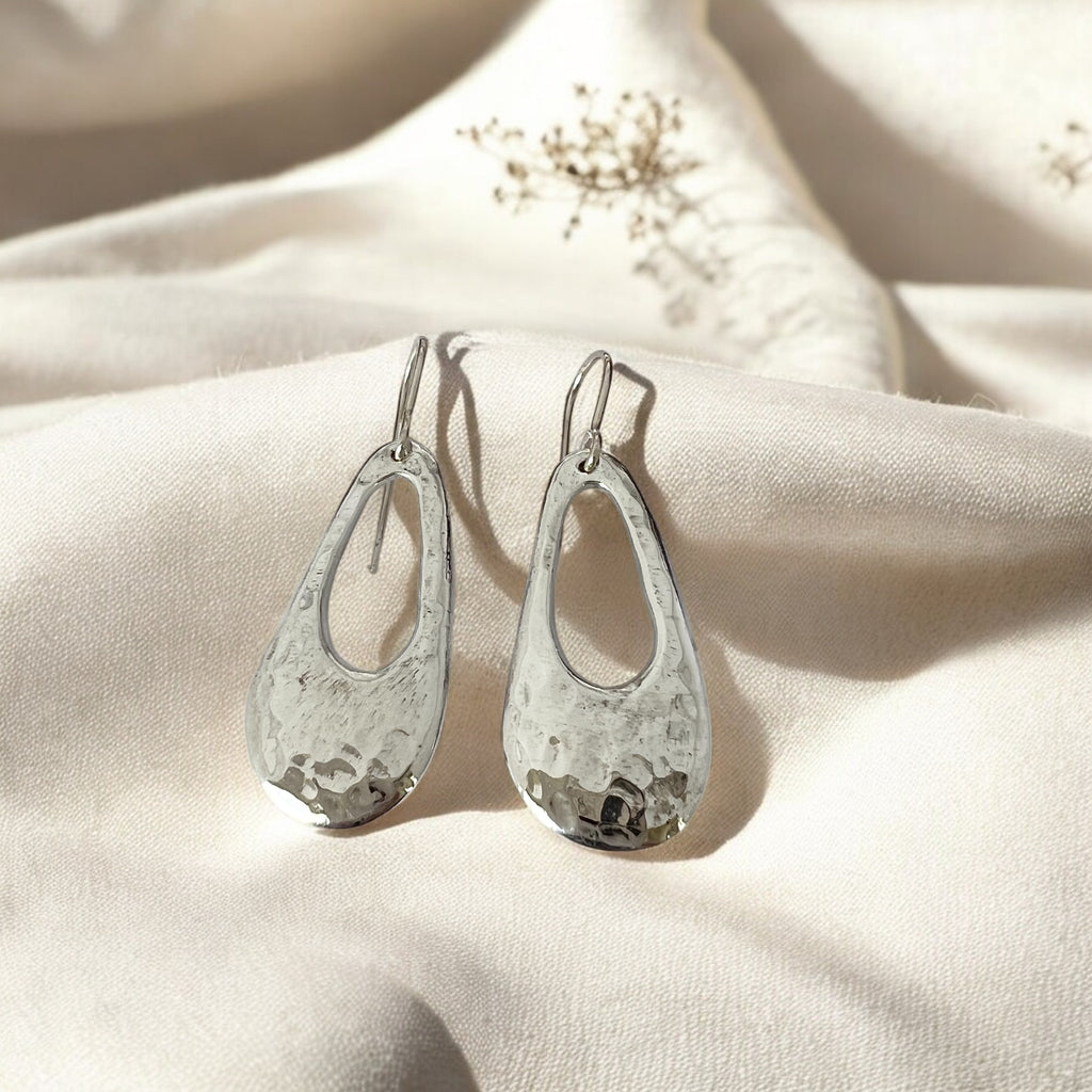 A pair of sterling silver freeform droplet earrings with a hammered texture, suspended from sterling silver hooks, displayed on a textured fabric.
