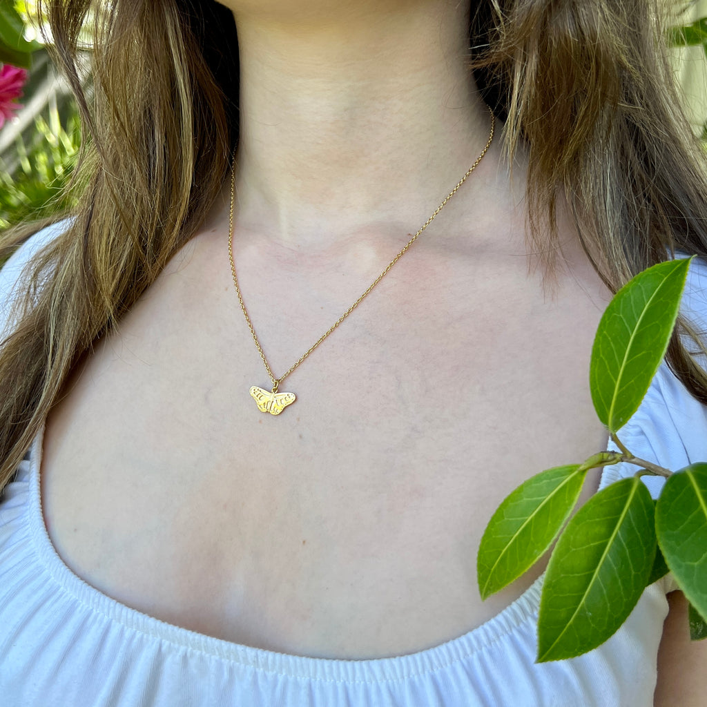 Person wearing a gold necklace with a butterfly pendant, holding green leaves.