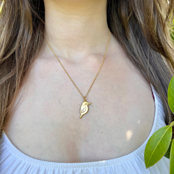 Gold necklace with a bird pendant worn by a person with greenery in the background