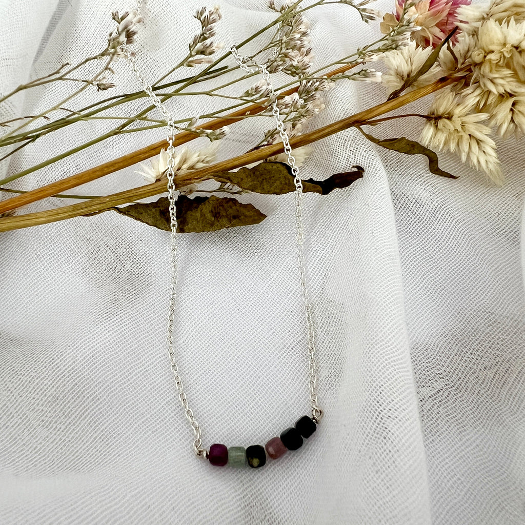 Silver ecklace with colored beads on a white fabric background with dried flowers.