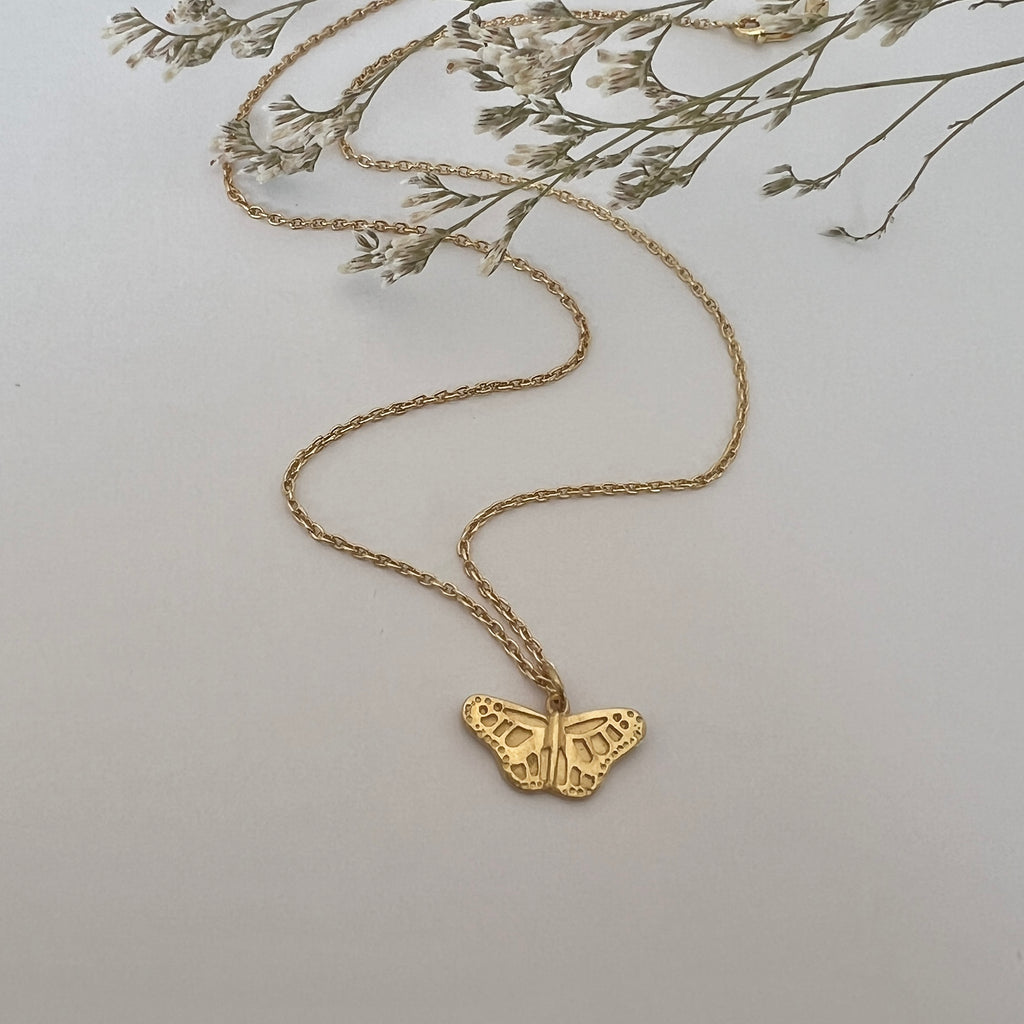 Gold butterfly necklace on a light gray background with dried flowers.