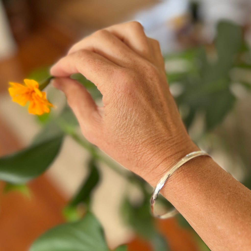A person's hand wearing a sterling silver bangle, holding a small yellow flower with a blurred background of greenery.