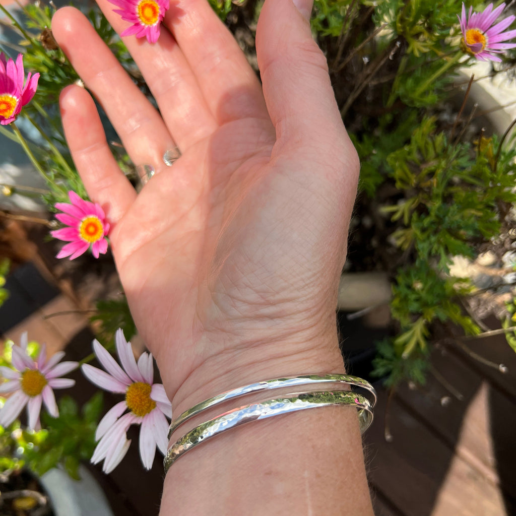 Hand with a silver bracelet and ring holding small pink flowers outdoors.