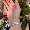 Hand with a silver bracelet and ring holding small pink flowers outdoors.