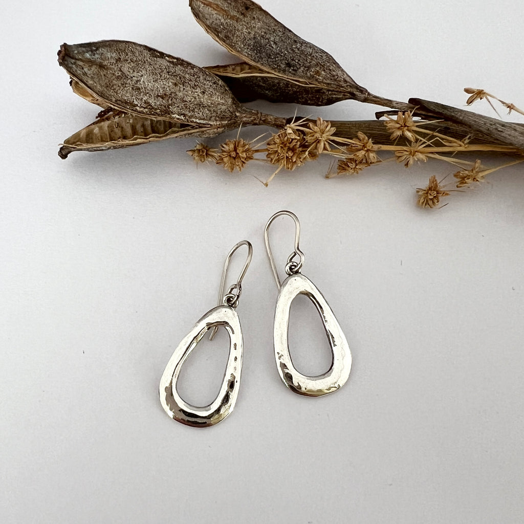 A pair of sterling silver earrings with a teardrop shape and a hammered texture, displayed against a white background with some dried plants in the background.