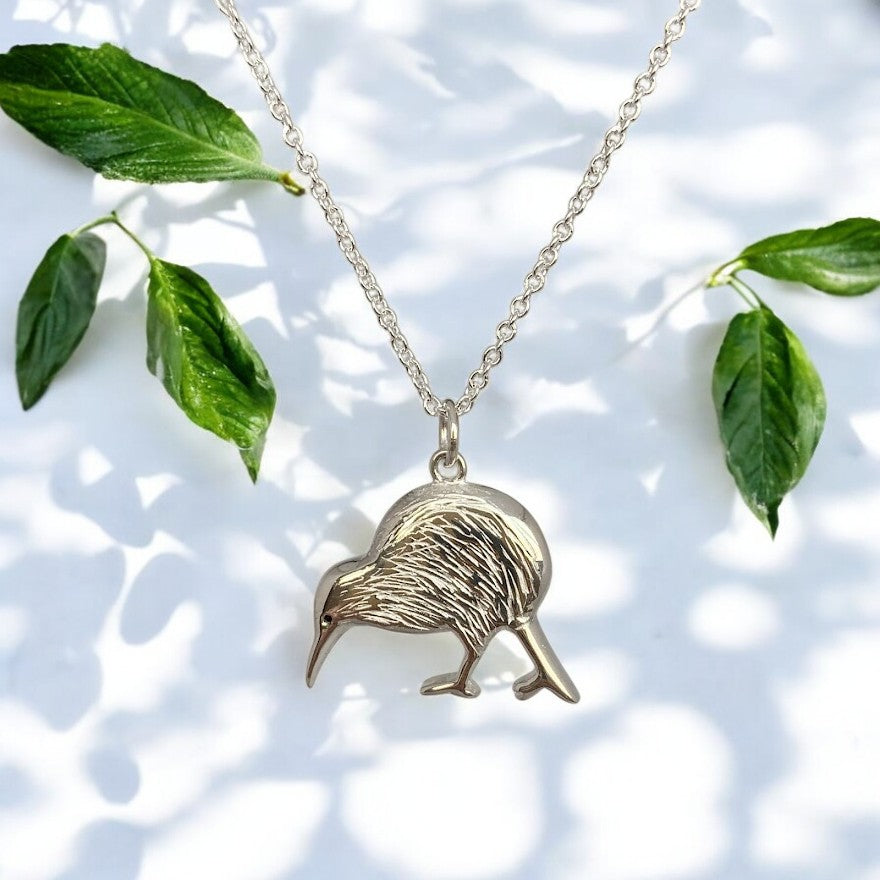 A sterling silver necklace featuring a detailed pendant of a Kiwi bird, accompanied by a sterling silver chain laid out on a patterned surface with green leaves.