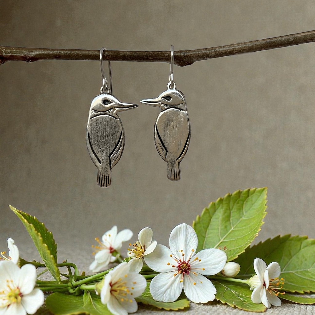 Silver bird-shaped earrings hanging from a branch with flowers and leaves on a textured surface.