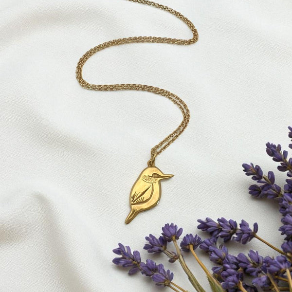 Gold necklace with a bird pendant on a white fabric background with lavender flowers.
