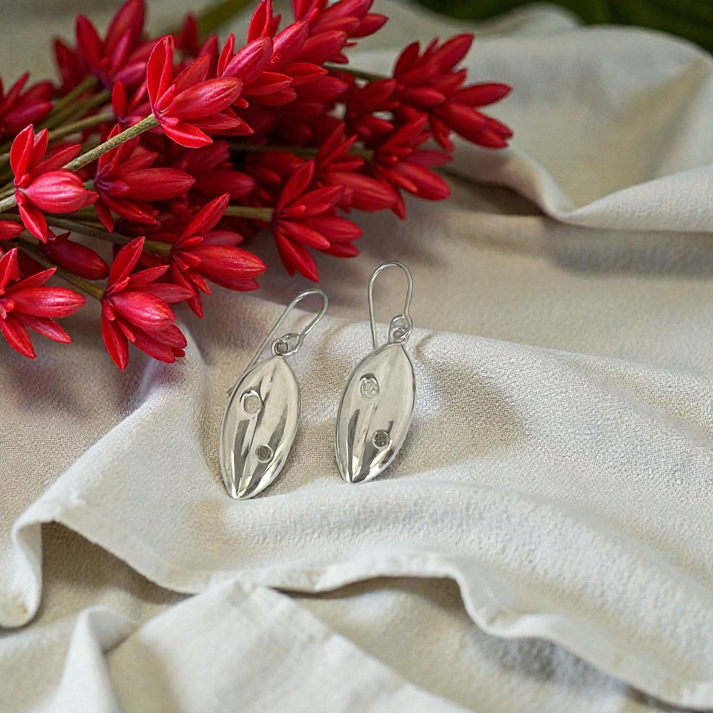 Silver Pohutukawa leaf earrings on a beige fabric with red flowers in the background