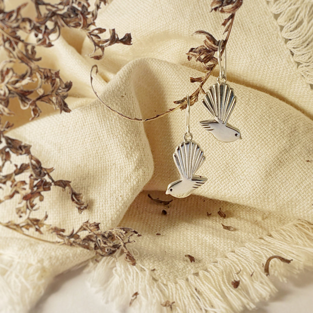 A pair of sterling silver pāwakawaka (Fantail) earrings, featuring the design of the bird in profile with its tail feathers fanned out, displayed against a textured beige fabric with some dried plant material.