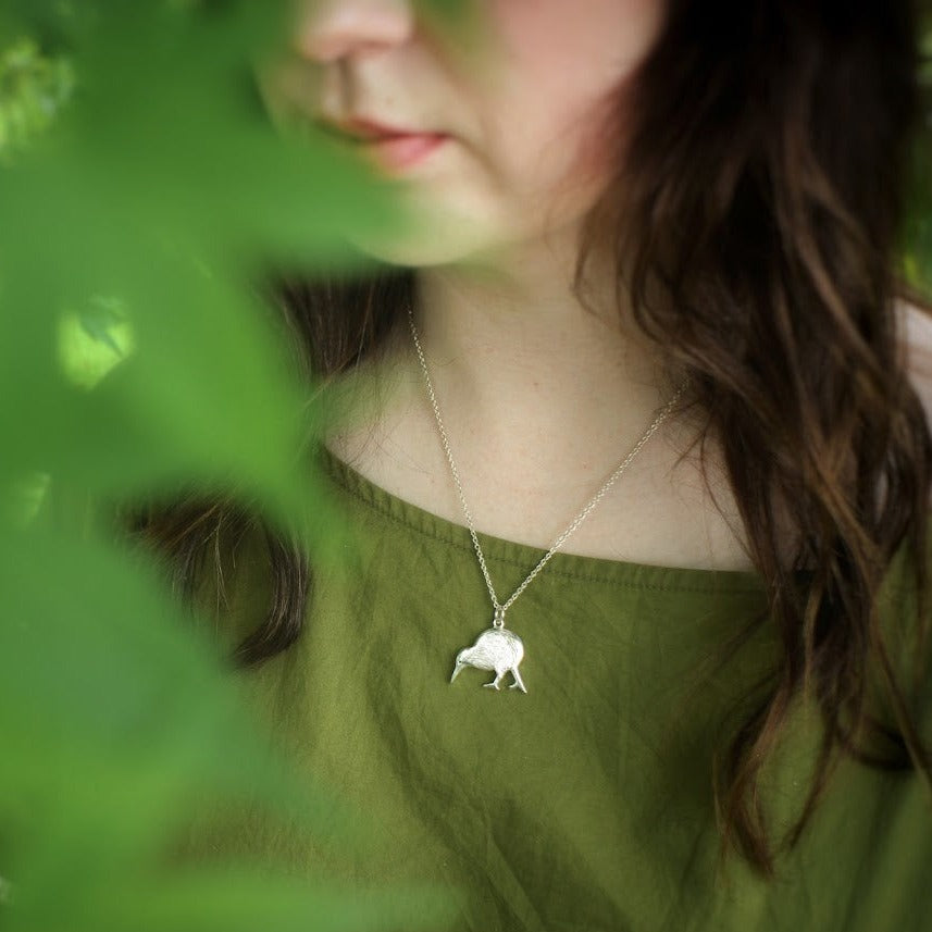 A person wearing a green shirt with a sterling silver necklace featuring a kiwi bird pendant, viewed through leaves.